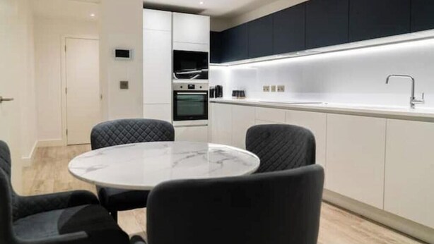 A modern kitchen with white cabinets, a black countertop, and a round dining table with chairs. - Modern 2-Bed Penthouse in Deansgate | Stunning City Views (Manchester)