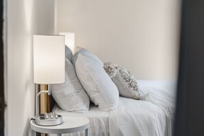 Een net opgemaakt bed met witte lakens en een nachtkastje met een lamp.