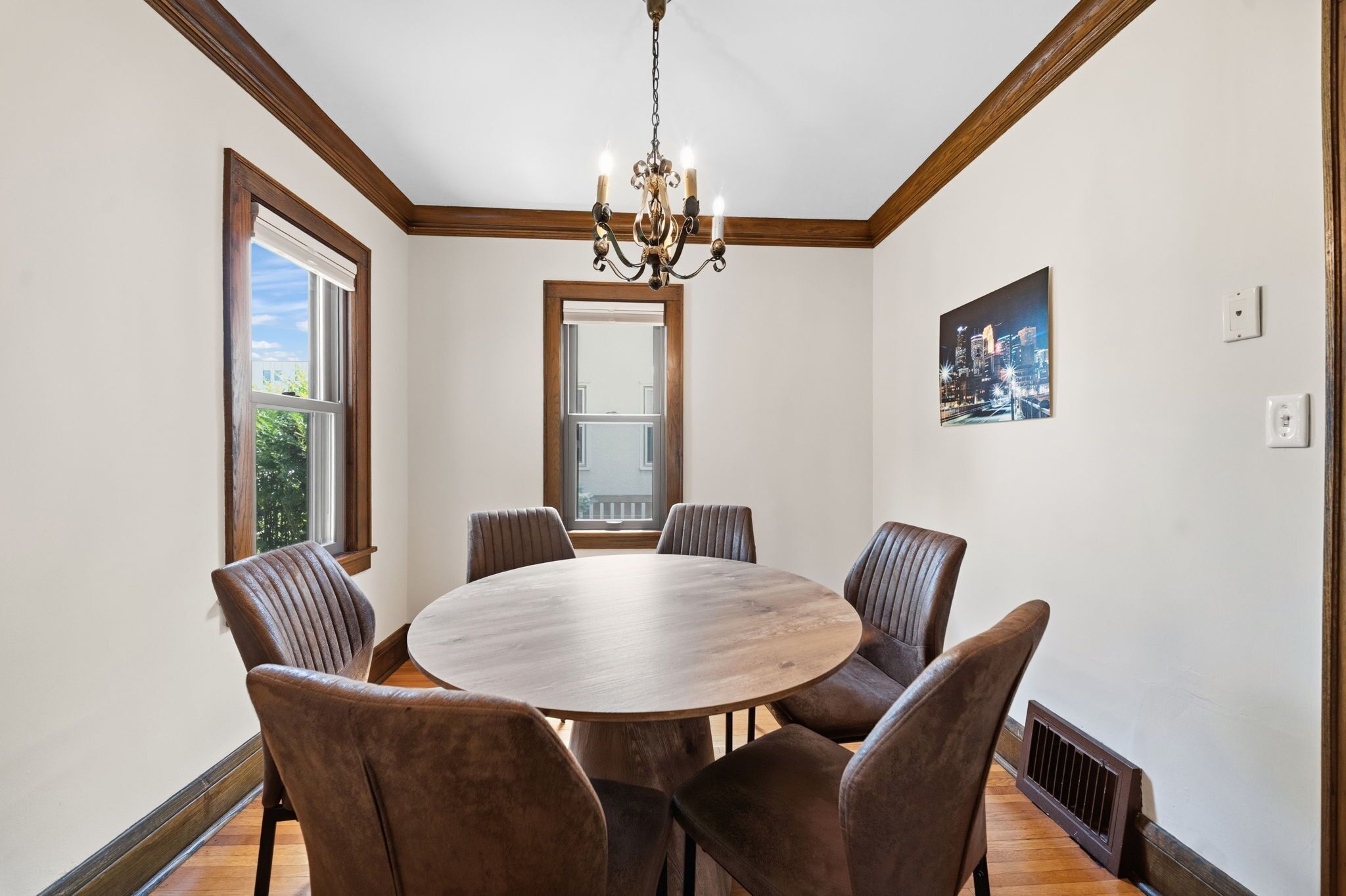 Ein Esszimmer mit einem runden Holztisch, sechs Polsterstühlen, einer Chandelierleuchtreibe, einem Stadtbildgemälde und einem großen Fenster.