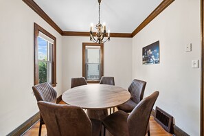 A dining room with a round wooden table, six upholstered chairs, a chandelier, a cityscape painting, and a large window. - MINNeSTAY* Magnolia House (Minneapolis)