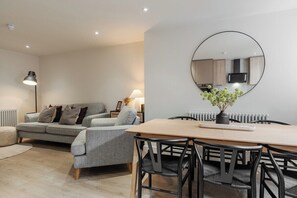Un salon moderne avec un canapé, une table à manger et des chaises. Un miroir rond est accroché au mur, et un radiateur se trouve à l’arrière-plan.
