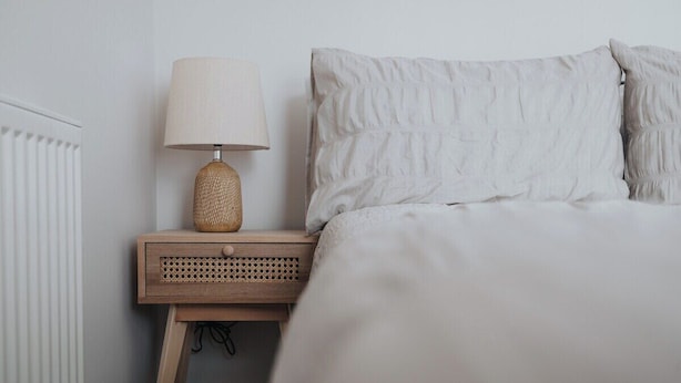 Ein ordentlich bezogenes Bett mit weißer Decke und Kissen, ein Nachttisch aus Holz mit einer gewobenen Lampe und ein weißer Heizkörper.
