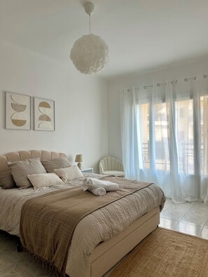 Ein Schlafzimmer mit einem Bett, Kissen, einem Nachttisch, einem Sessel und einem Fenster mit Vorhängen.