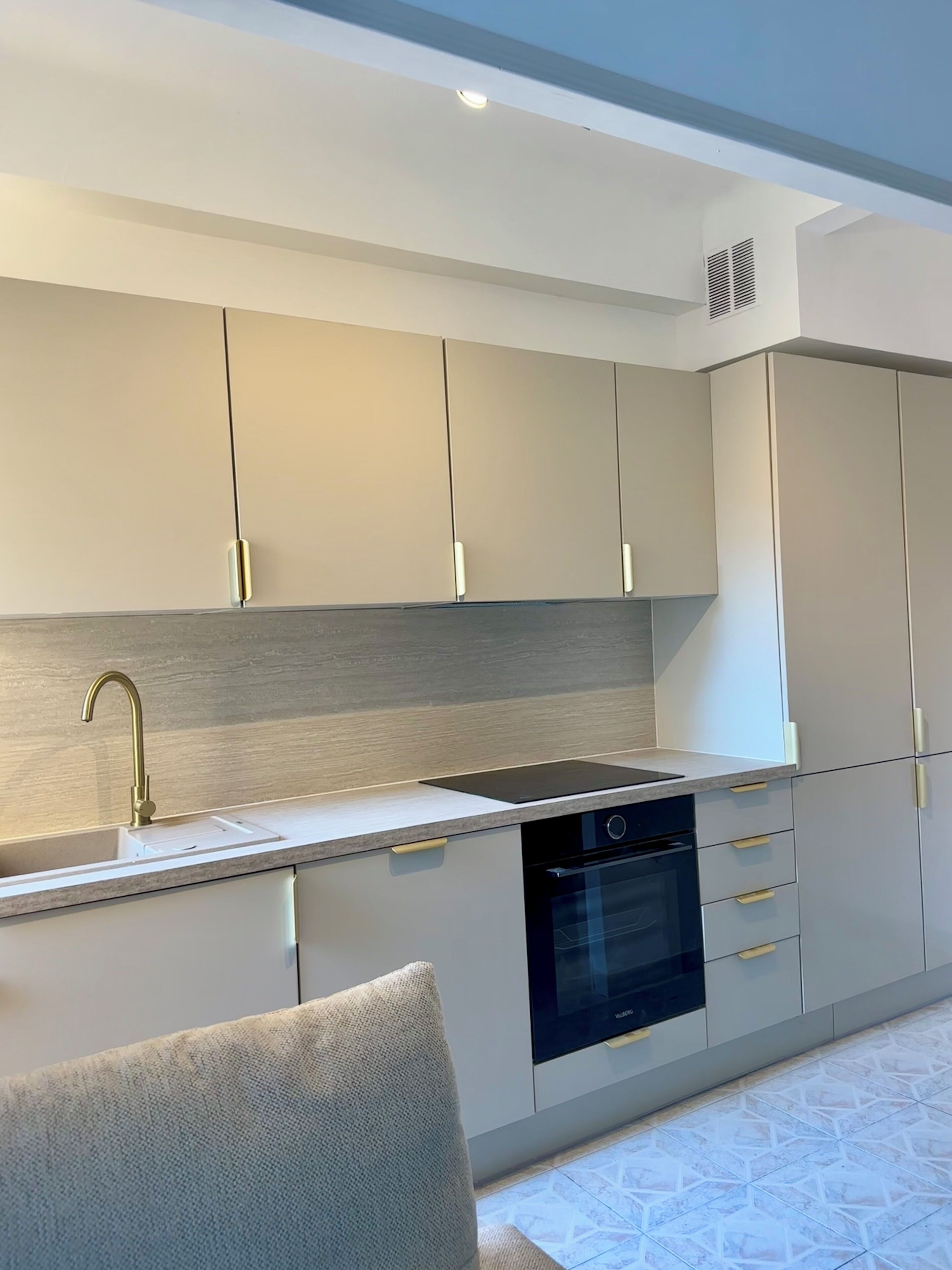A modern kitchen with a built-in oven, a sink with a gold faucet, and light-colored cabinets.