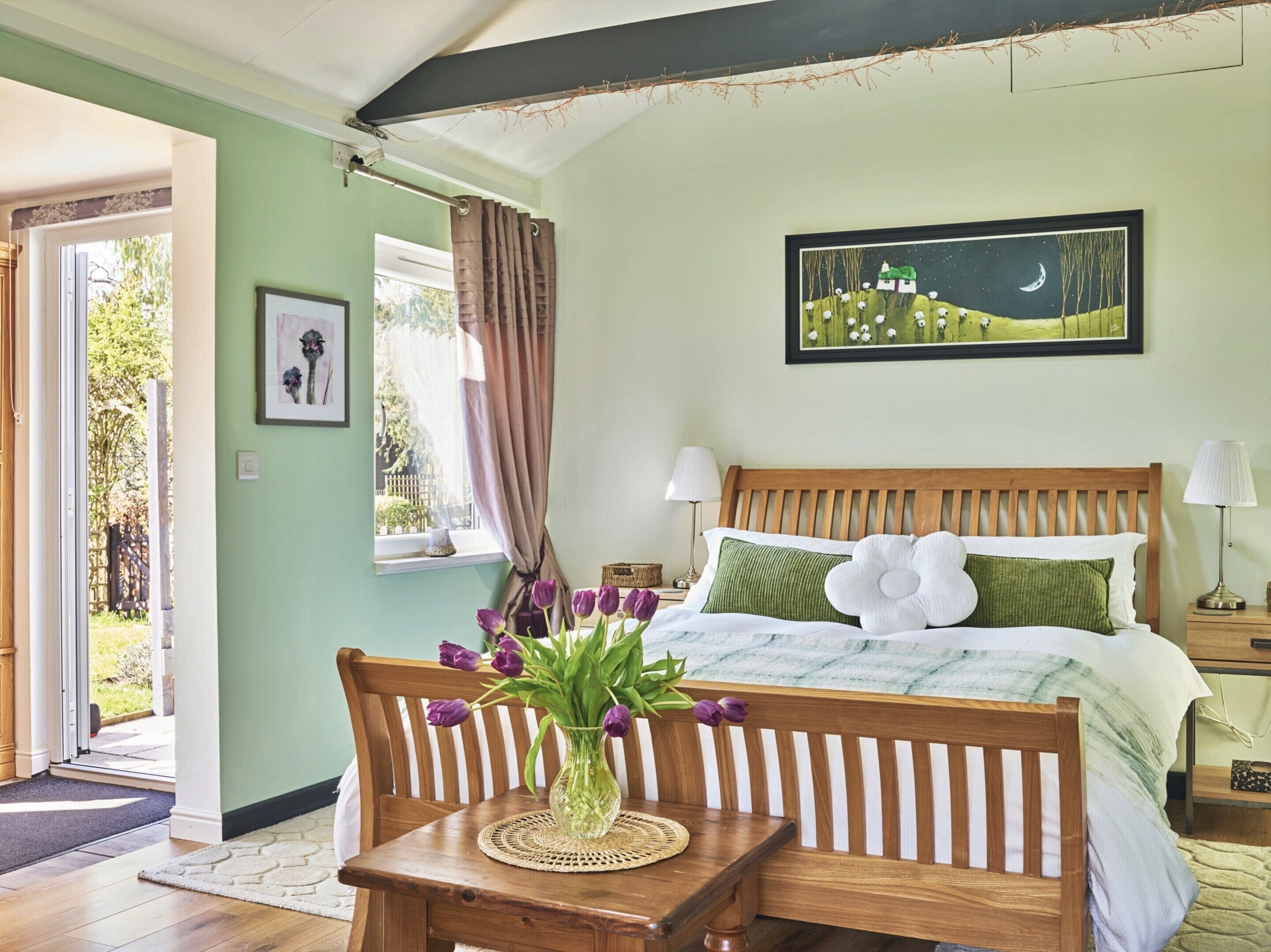Une chambre à coucher avec un lit en bois, une table de chevet, une petite table de nuit ornée d’un vase de fleurs et une fenêtre avec des rideaux.