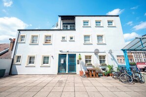 A two-story building with a blue door, multiple windows, and a tiled patio area with potted plants and bicycles. - Comfortable Stay in the Heart of Vibrant Deptford - Pass the Keys (London)