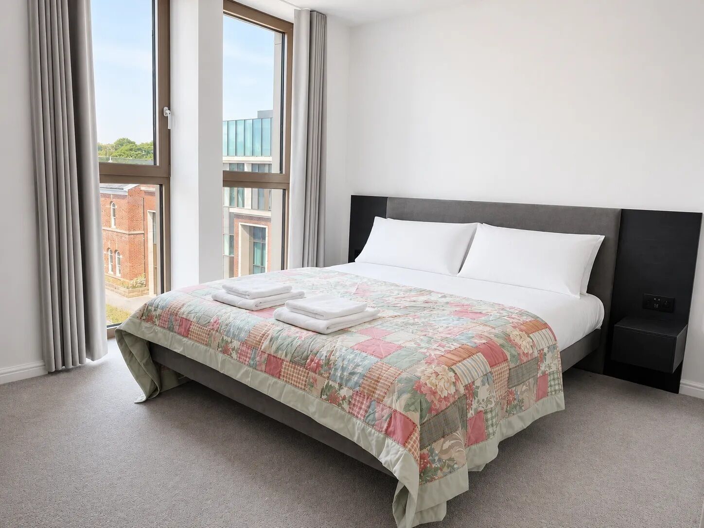 A neatly made bed with a floral quilt, a window with curtains, and a view of buildings outside.