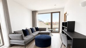 Ein modernes Wohnzimmer mit einem grauen Ecksofa, einem runden Hocker, einem kleinen runden Tisch, einem schwarzen Schreibtisch mit Fernseher und großen Fenstern mit Blick auf Gebäude.