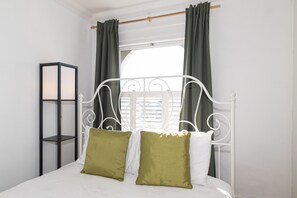 Ein Bett mit Metallgestell, zwei Kissen, ein Nachttisch und ein Fenster mit Vorhängen.