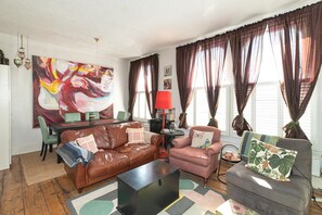 Un soggiorno con un divano in pelle marrone, una poltrona rosa, un tavolino da caffè e un divano grigio con un cuscino floreale. C'è un quadro appeso al muro, una lampada rossa su un comodino e una finestra con tende.