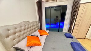 Een slaapkamer met een gestoffeerd hoofdeinde, een grijze spreideken, twee oranje kussens en een blauwe handdoek.