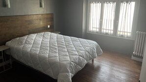 Quarto com cama grande, cabeceira de madeira, janela com cortinas leves e radiador.
