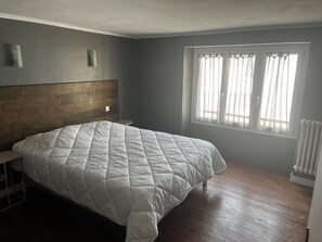 Ein Schlafzimmer mit einem großen Bett, einem Holz-Kopfteil, einem Fenster mit transparenten Vorhängen und einem Heizkörper.