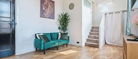A room with a teal sofa, a plant, a staircase, and a TV on a wooden stand.