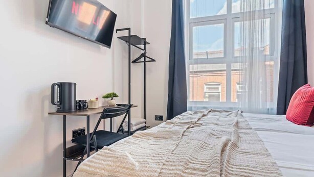 Ein Hotelzimmer mit einem Bett, einem an der Wand befestigten Fernseher, einem Schreibtisch mit Wasserkocher und einem Fenster mit Blick auf Gebäude.
