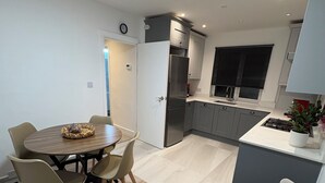 A modern kitchen with a built-in oven and refrigerator, a dining area with a round table and chairs, and a living area with a sofa and a potted plant. - Modern 3 bedroom Apartment, Sleep 5 (Barnet)