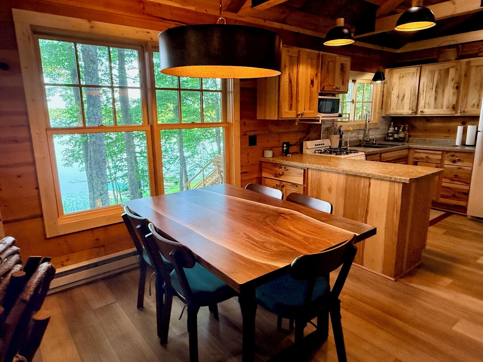 Interior de una cabaña de madera con una mesa de comedor, sillas, una zona de cocina con microondas y un ventanal que ofrece vistas a árboles.