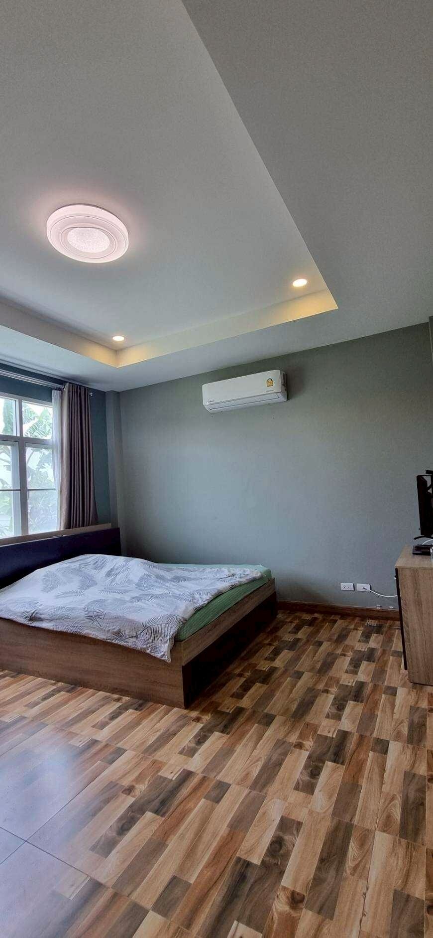 A bedroom with a wooden floor, a bed, a window with curtains, an air conditioner, and a desk with a computer.