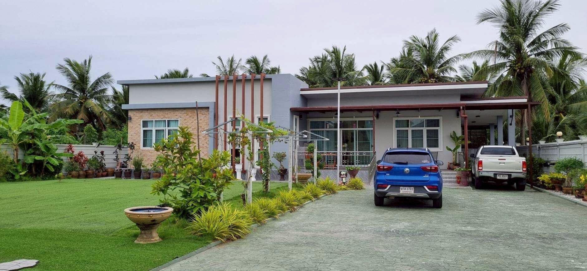 A modern two-story house with a paved driveway, parked vehicles, and a well-maintained garden with palm trees and various plants.