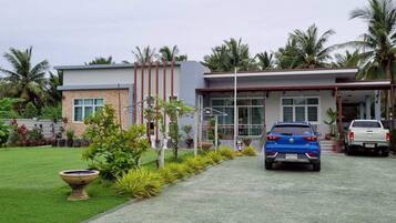 A modern two-story house with a paved driveway, parked vehicles, and a well-maintained garden with palm trees and various plants.