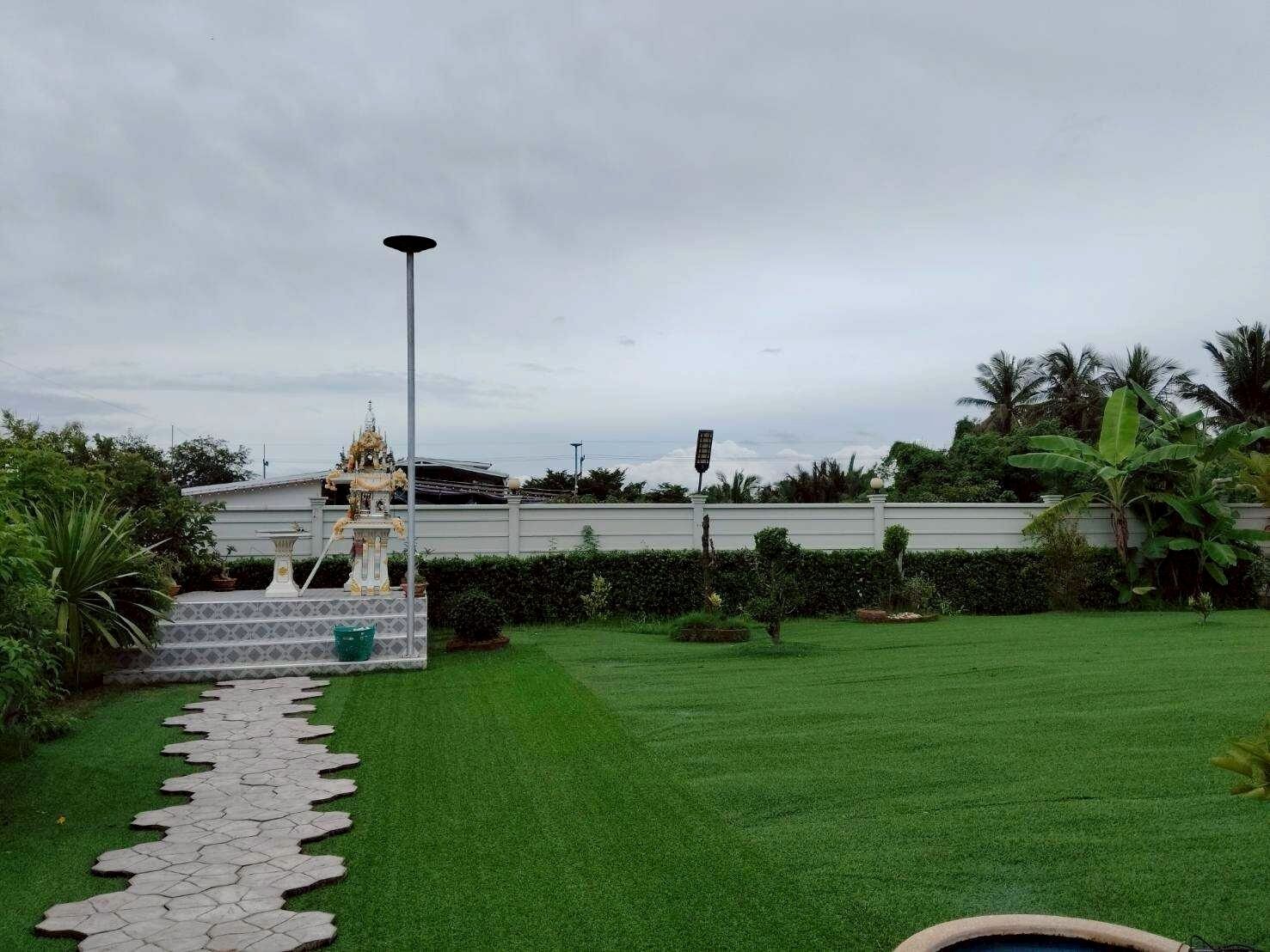 Un jardin bien entretenu, avec un chemin de pierre menant à une fontaine décorative, entouré d’une végétation luxuriante et, au fond, d’un bâtiment blanc.