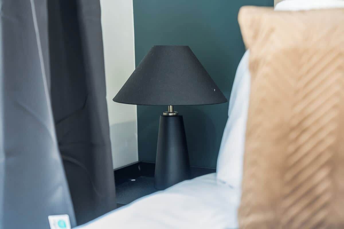 Ein Bett mit einer schwarzen Lampe daneben, einem braunen Kopfkissen und einem dunklen Vorhang, der das Fenster teilweise verdeckt.