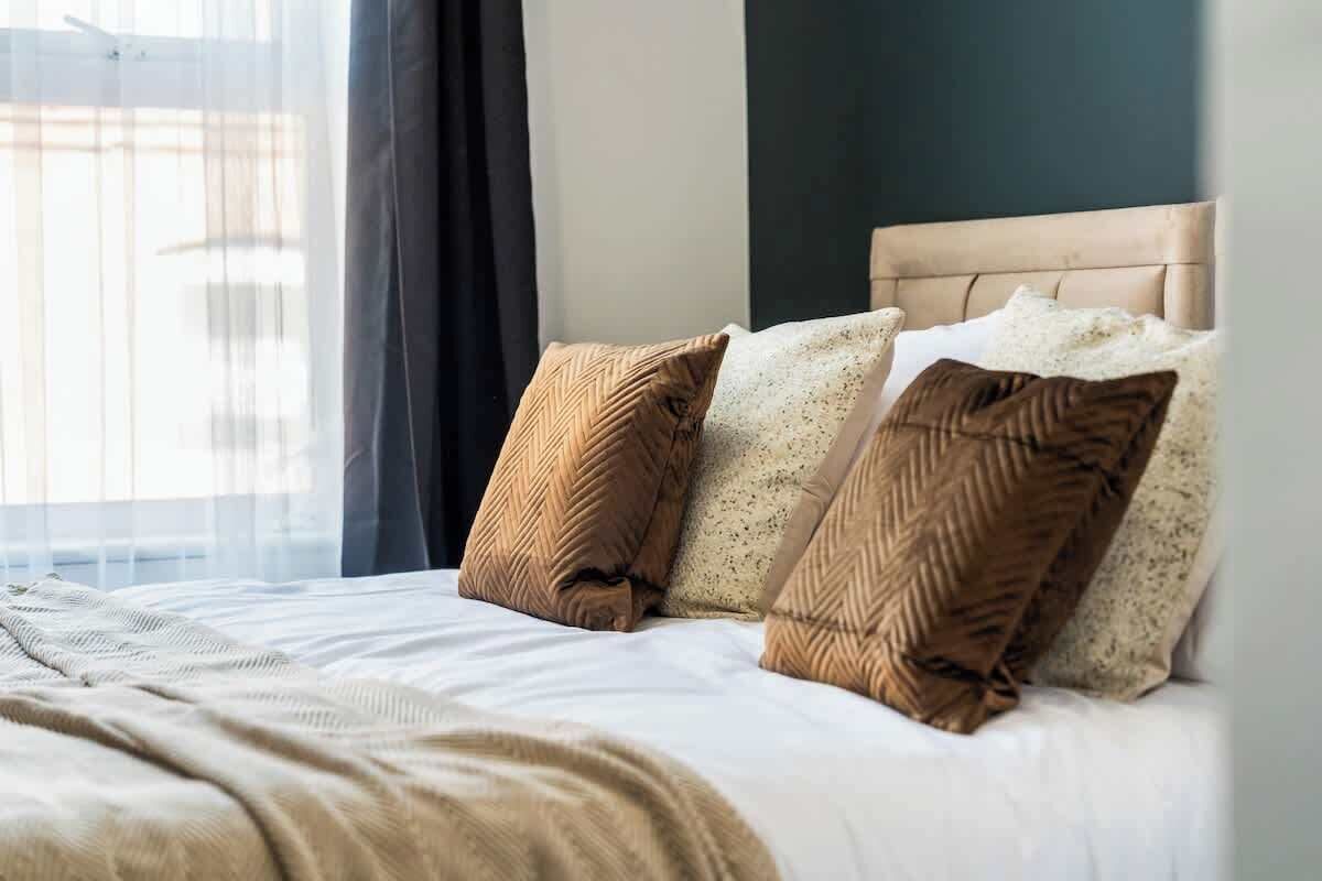 A neatly made bed with a headboard, a window with sheer curtains, and a view of the outside.