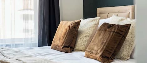 A neatly made bed with a headboard, a window with sheer curtains, and a view of the outside.