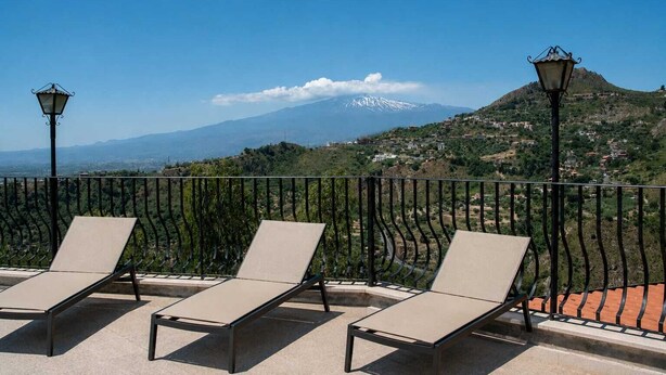 A terrace with three sun loungers, a metal railing, and a view of a mountain with a snow-capped peak.