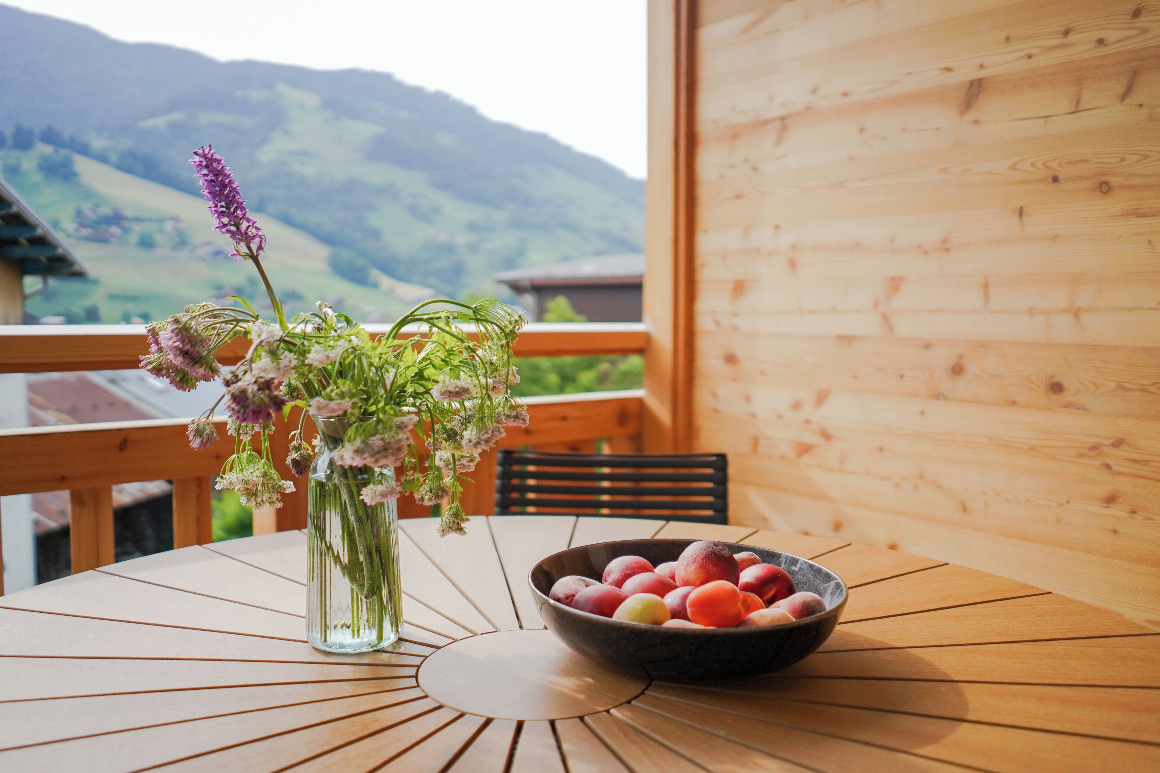 Uma mesa com um vaso de flores e uma fruteira numa varanda com vista para as montanhas.