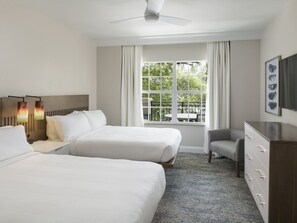 Habitación de hotel con dos camas, un escritorio, una silla, un aparador, un ventilador de techo y una ventana con vistas a zonas verdes.