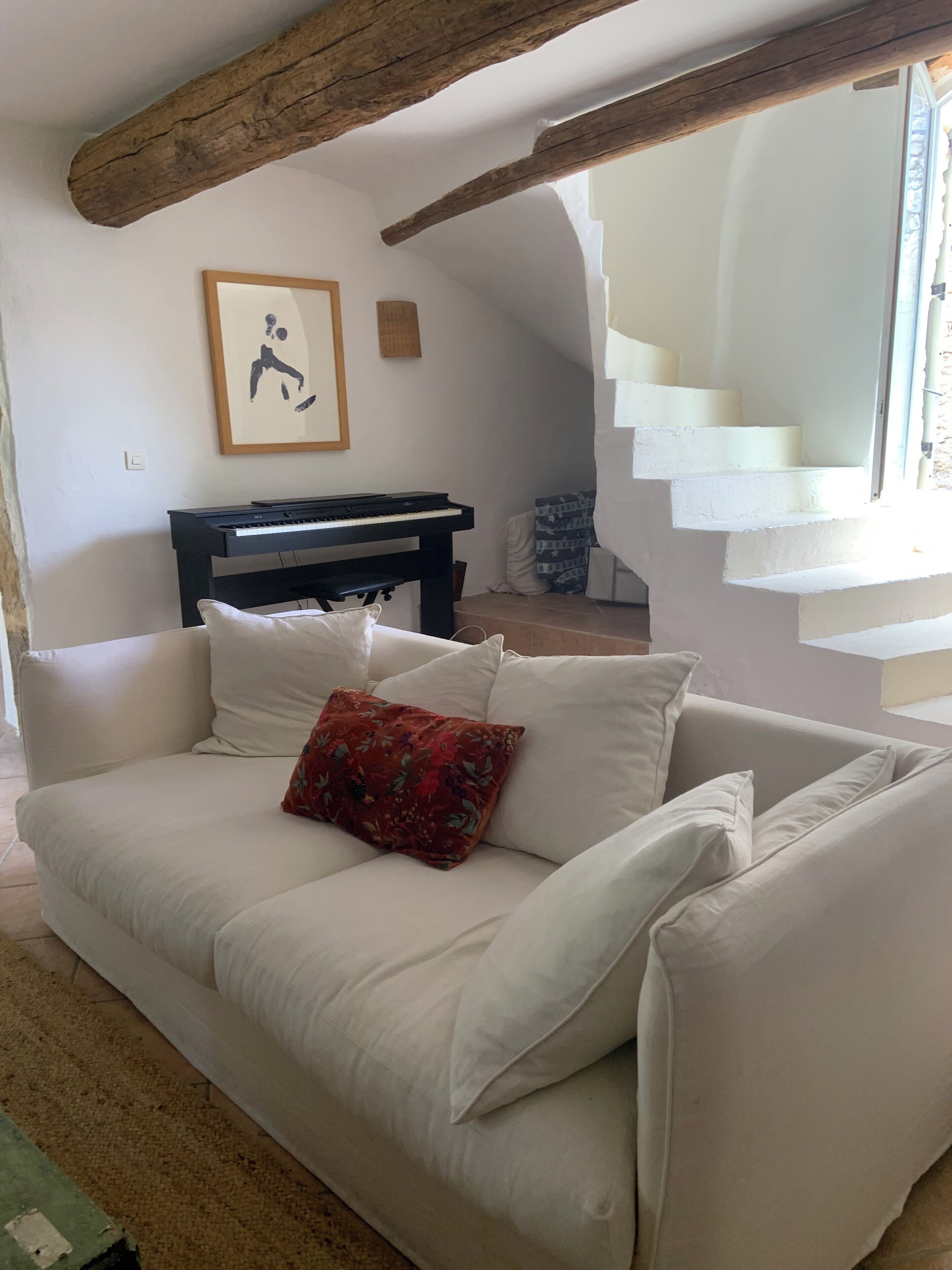 A living room with a white sofa, a red floral pillow, a black piano, and a staircase leading to an upper level.