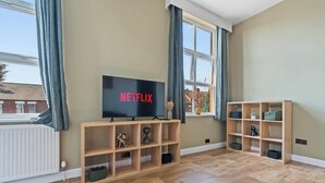 Ein Wohnzimmer mit Fernseher, der das Netflix-Logo zeigt, Holzregalen und einem großen Fenster mit Vorhängen.