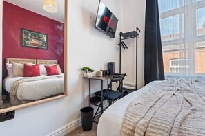 A hotel room with a bed, a desk with a chair, a TV mounted on the wall, and a window with a view of buildings. - Central Studio with Double Bed | CV2 |Free Parking (Coventry)