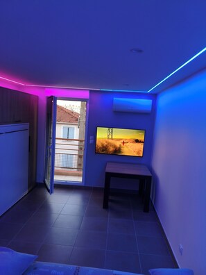 A room with a large flat-screen TV mounted on the wall, a bench, and a sliding door leading to an outdoor area. - The Albino Oracle (Nîmes)