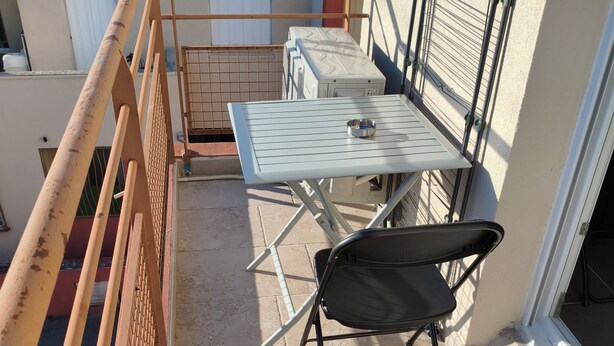 A balcony with a metal table and chair, a metal railing, and a view of a building. - The Albino Oracle (Nîmes)