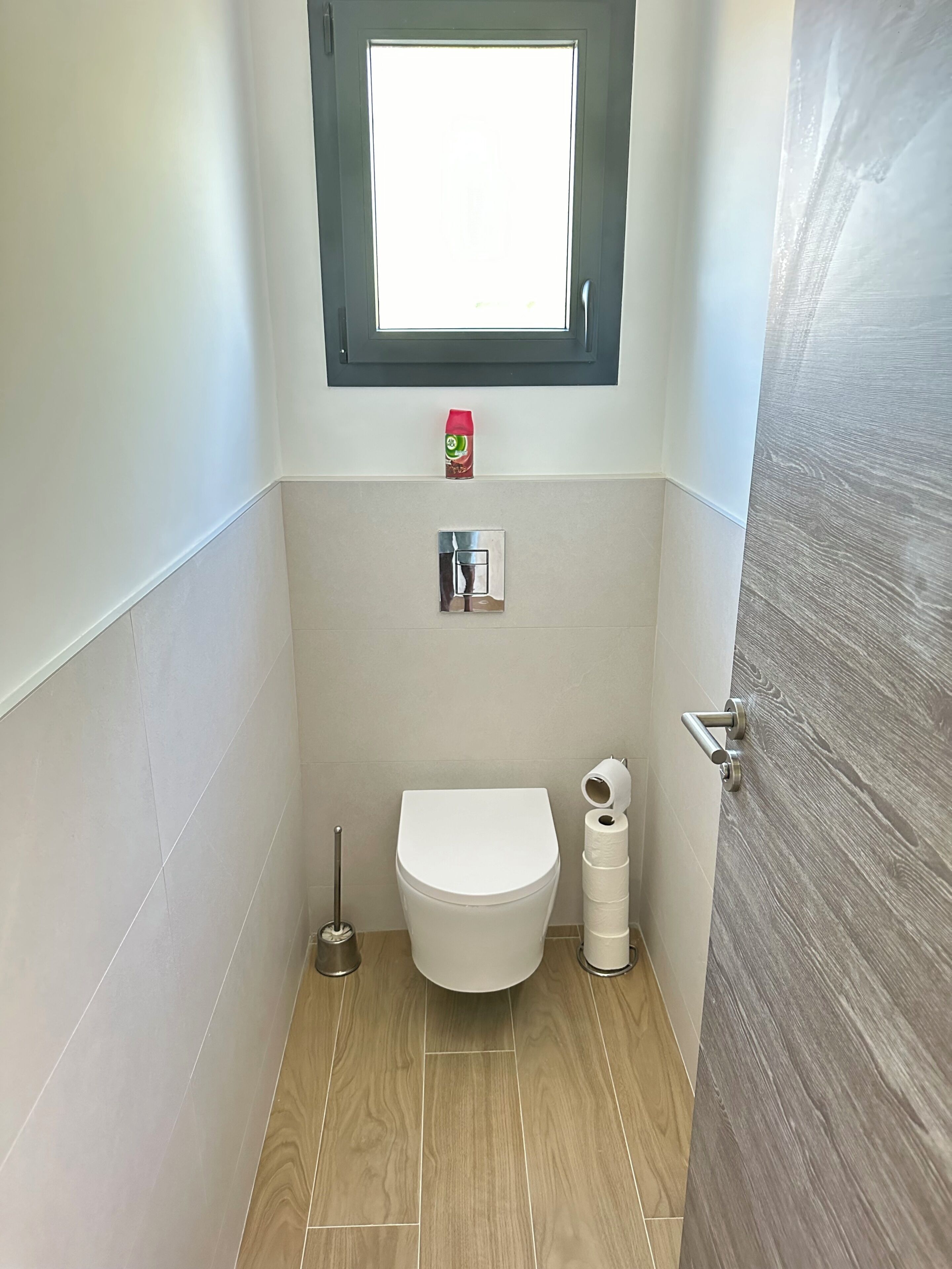 Een kleine badkamer met toilet, een douchetoilet, een toiletrolhouder, een raam en een deur.