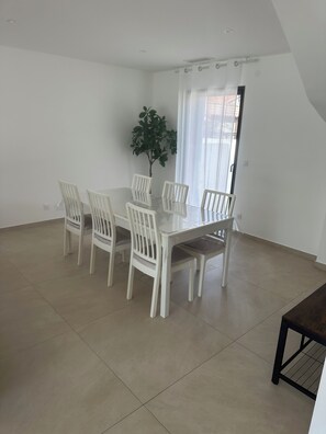 A dining area with a white table and chairs, a potted plant, and a view of the outside through a window with curtains. - Large, air-conditioned family home with a pool in Toulon, Var (La Valette-du-Var)