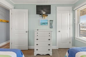 Ein Schlafzimmer mit zwei weißen Türen, einer Kommode mit Schubladen, einem Wand-Fernseher und einem Fenster mit Jalousien.