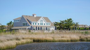 A large house with a prominent gable roof, a fenced deck, and a body of water in the foreground. - Making Memories: Oceanfront 6 BR | Hot Tub | Shared Pool (Duck)