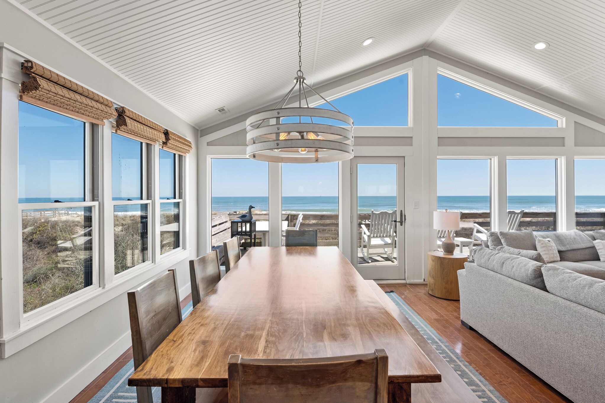 A spacious room with a large wooden dining table, a gray sofa, and multiple windows offering ocean views.