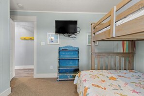 A bedroom with a bunk bed, a television mounted on the wall, a blue dresser, and a door leading to another room. - Making Memories: Oceanfront 6 BR | Hot Tub | Shared Pool (Duck)