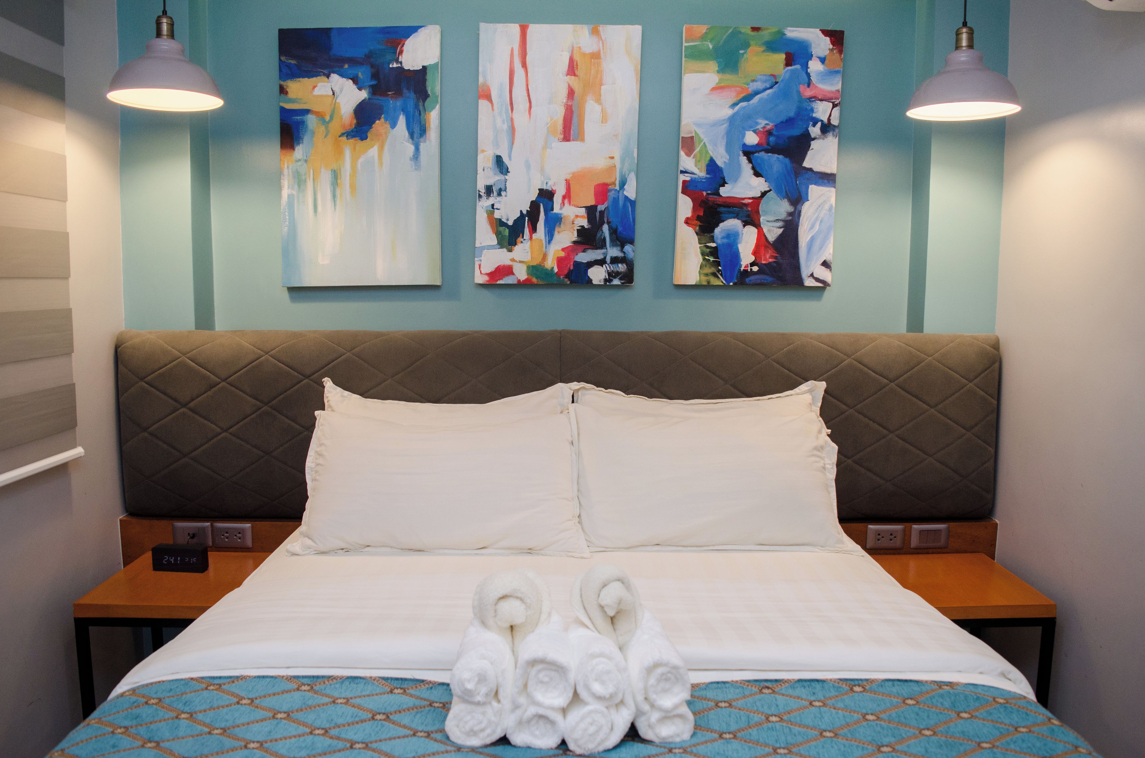 Una habitación de hotel con cama, dos mesitas de noche, una pared con pinturas abstractas y dos lámparas colgantes.