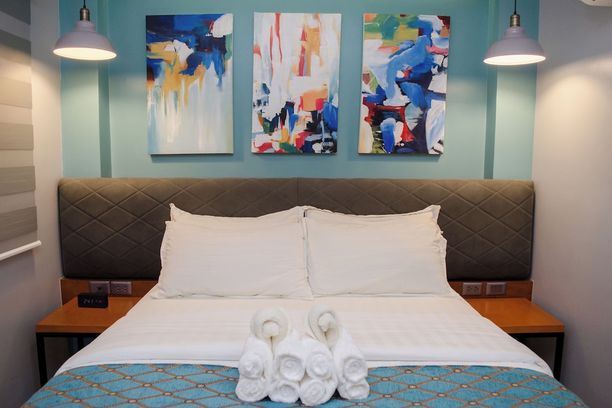 A hotel room with a bed, two bedside tables, a wall with abstract paintings, and two pendant lights.