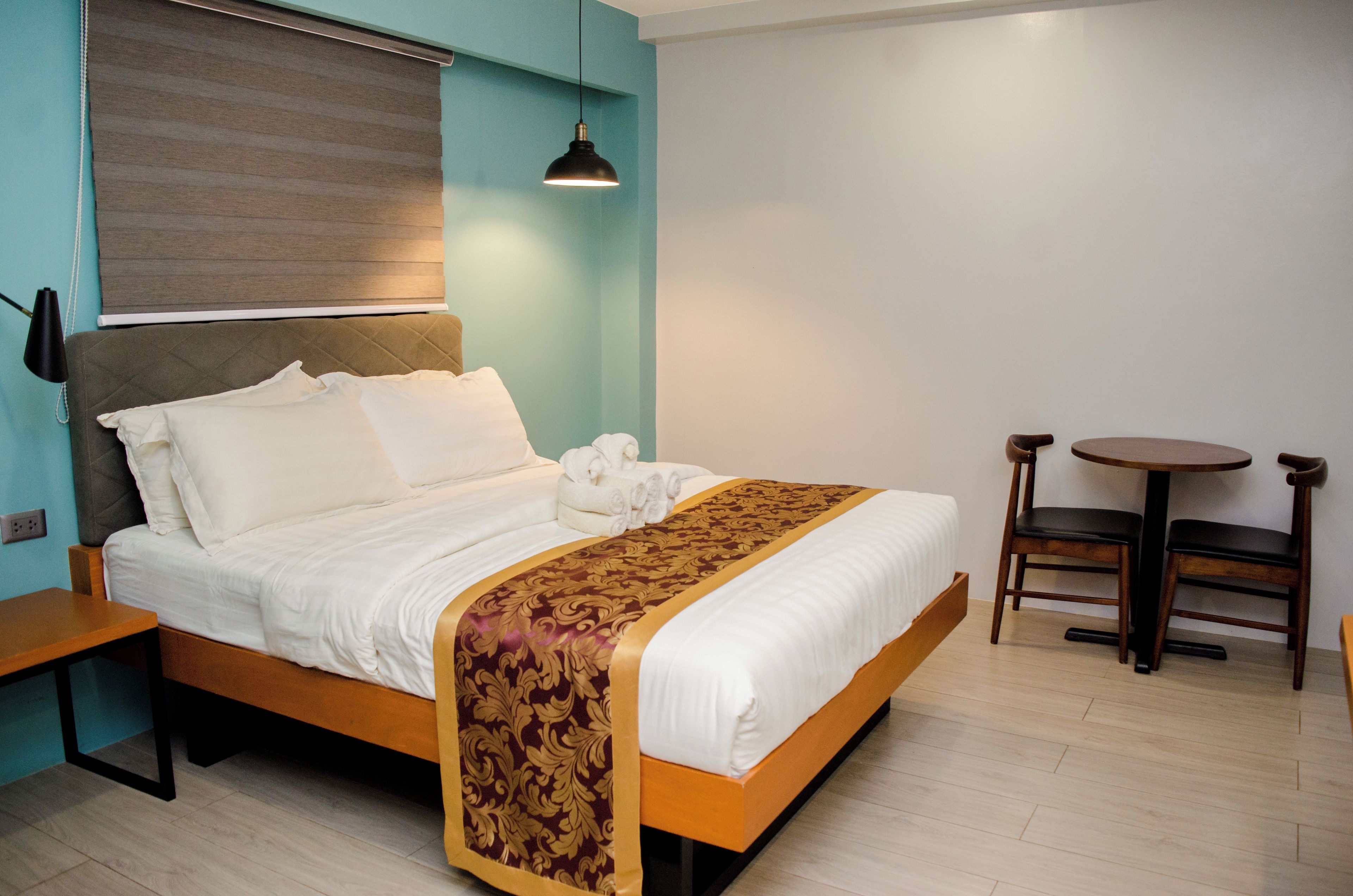 A neatly made bed with a patterned bedspread, a bedside table, a lamp, and a small round table with two chairs.