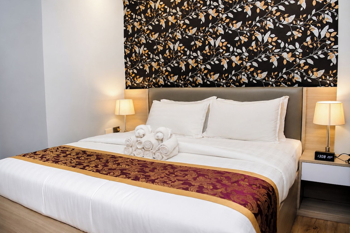 Una habitación de hotel con una cama, dos lámparas de noche, un reloj digital y un adorno en la pared sobre la cama.