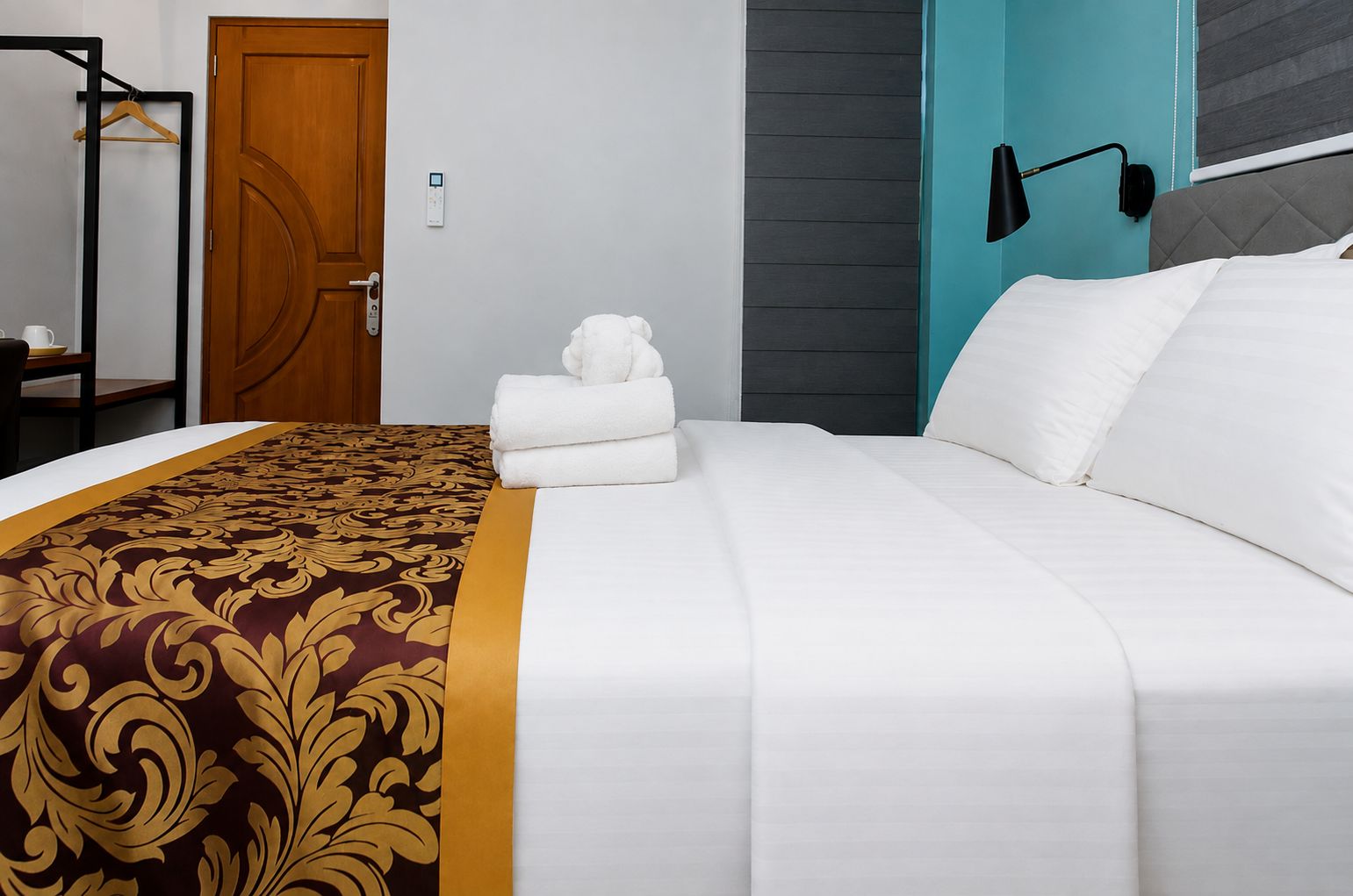 A hotel room with a bed, a wooden door, a coat rack, and a wall-mounted light fixture.