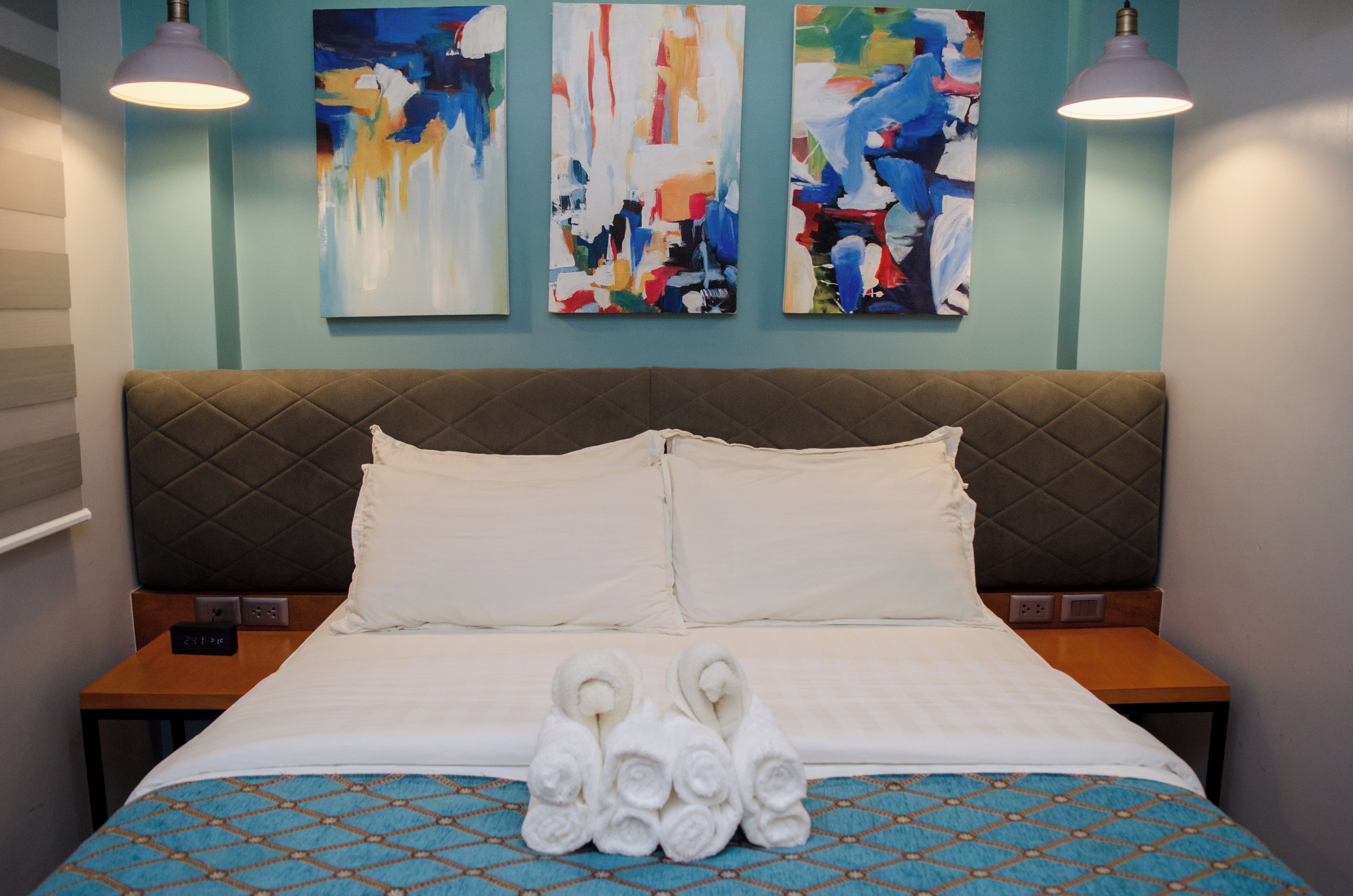 A hotel room with a bed, two bedside tables, a headboard, and three abstract paintings on the wall.