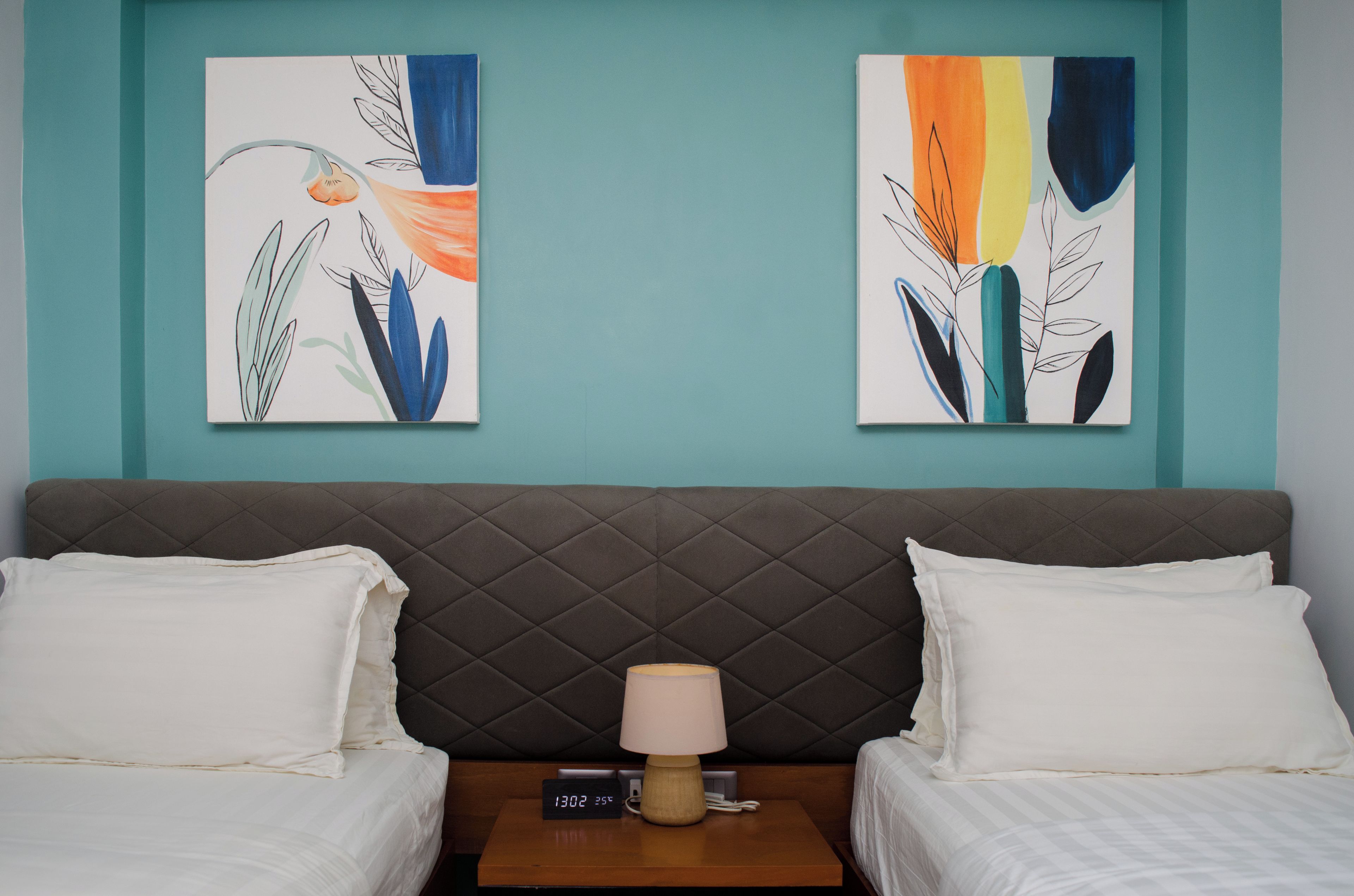 A hotel room with a bed, two white pillows, a nightstand with a lamp, and two abstract paintings on the wall.