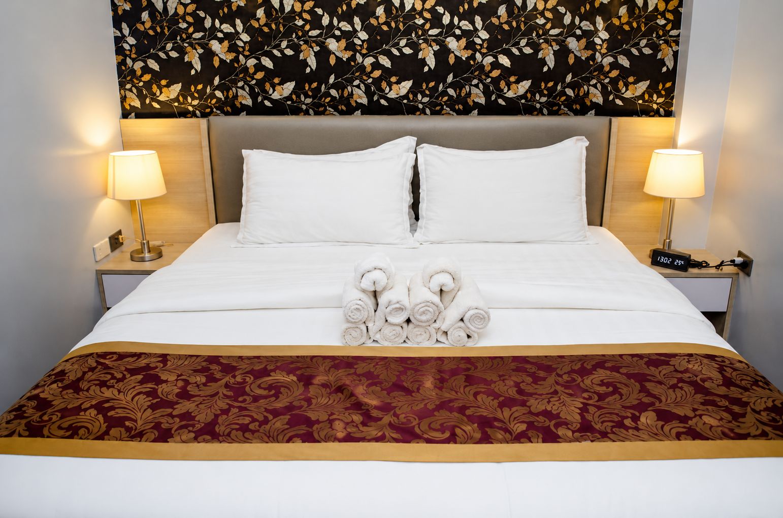 Habitación de hotel con una cama, dos lámparas de noche, un reloj digital y una pared decorativa con un diseño de hojas.